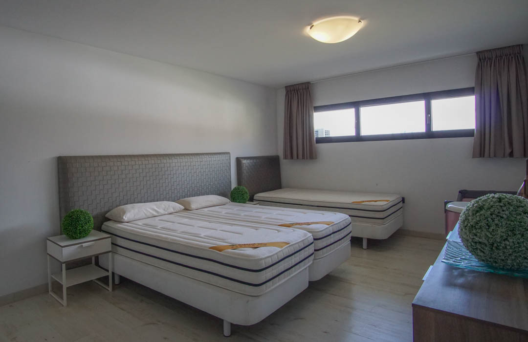 Bedroom featuring two single beds with grey patterned headboards, a third single bed in the background, light wood flooring, and a series of horizontal windows.