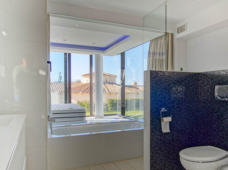 Contemporary bathroom with a built-in bathtub, floor-to-ceiling windows offering views of a tiled roof and garden. Features blue accent lighting and a dark patterned partition.