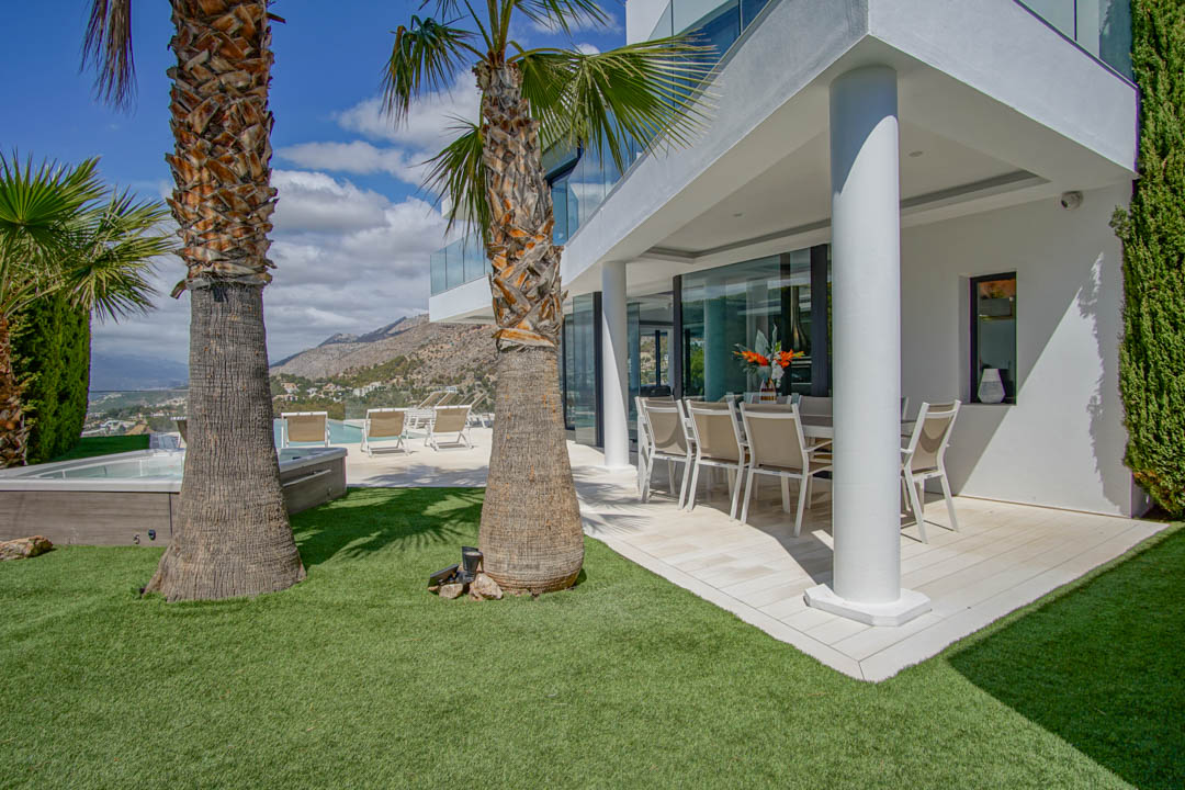 Covered outdoor dining terrace with modern furniture, adjacent to a lush artificial lawn and prominent palm trees. A jacuzzi and pool area offer mountain and distant sea views.