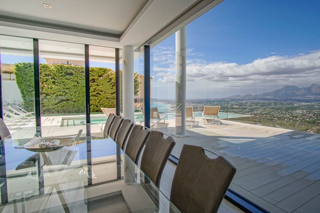 Elegant indoor dining area featuring a glass table and chairs, seamlessly connecting to an expansive terrace with an infinity pool and panoramic sea and mountain vistas.