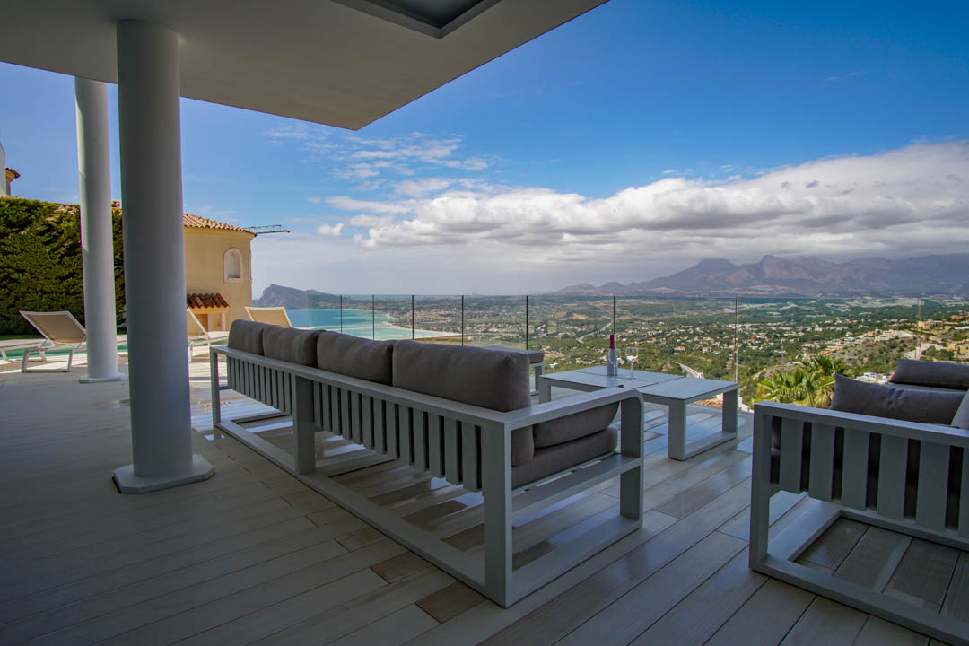 Expansive covered terrace with light decking and modern white lounge furniture, framing panoramic sea, coastal, and mountain views through a glass railing.