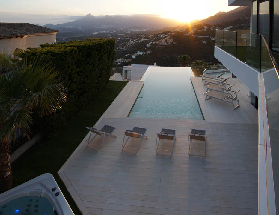 Expansive infinity pool with a light-toned deck, multiple sun loungers, and a separate jacuzzi, all set against a panoramic mountain sunset backdrop.