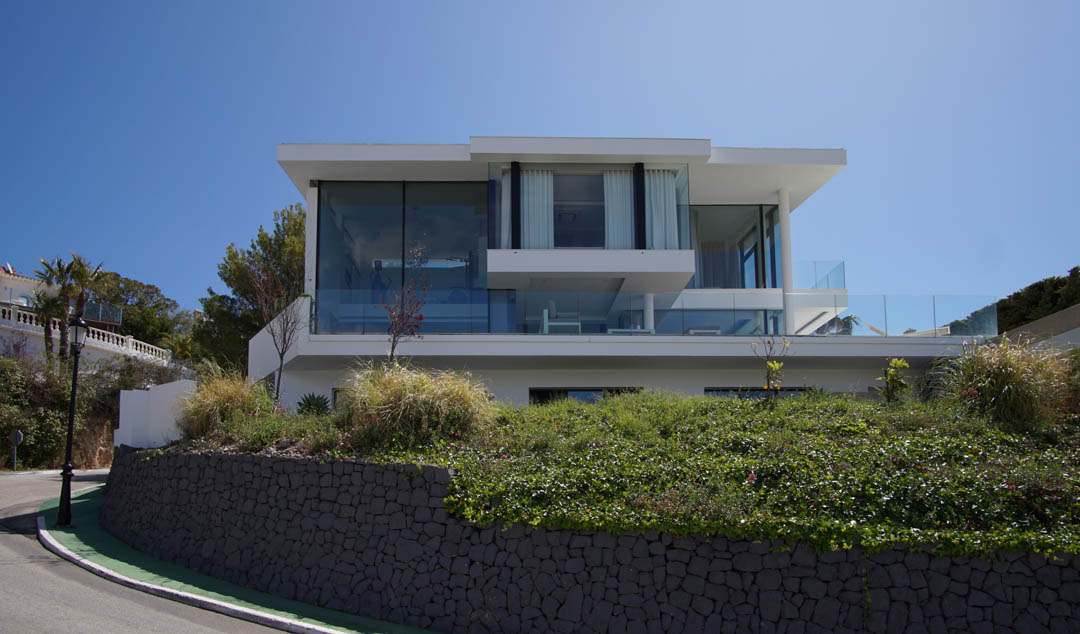 Exterior view of a modern three-level villa featuring extensive glass facades and glass railings, elevated above a stone retaining wall and lush vegetation by a curved road.