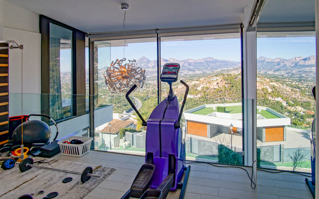 Gym with floor-to-ceiling windows showcasing panoramic mountain and valley views. Features an elliptical machine, free weights, and a glass balustrade in a modern setting.
