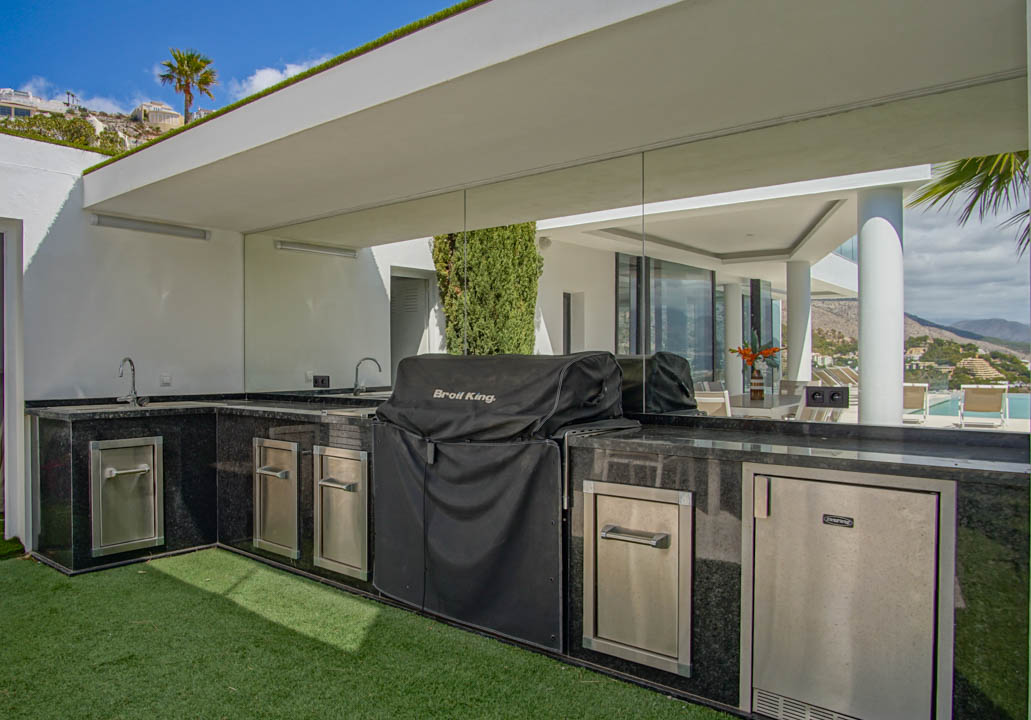 L-shaped outdoor kitchen featuring black granite countertops, dual sinks, a covered BBQ grill, and integrated stainless steel appliances, with reflected pool views.