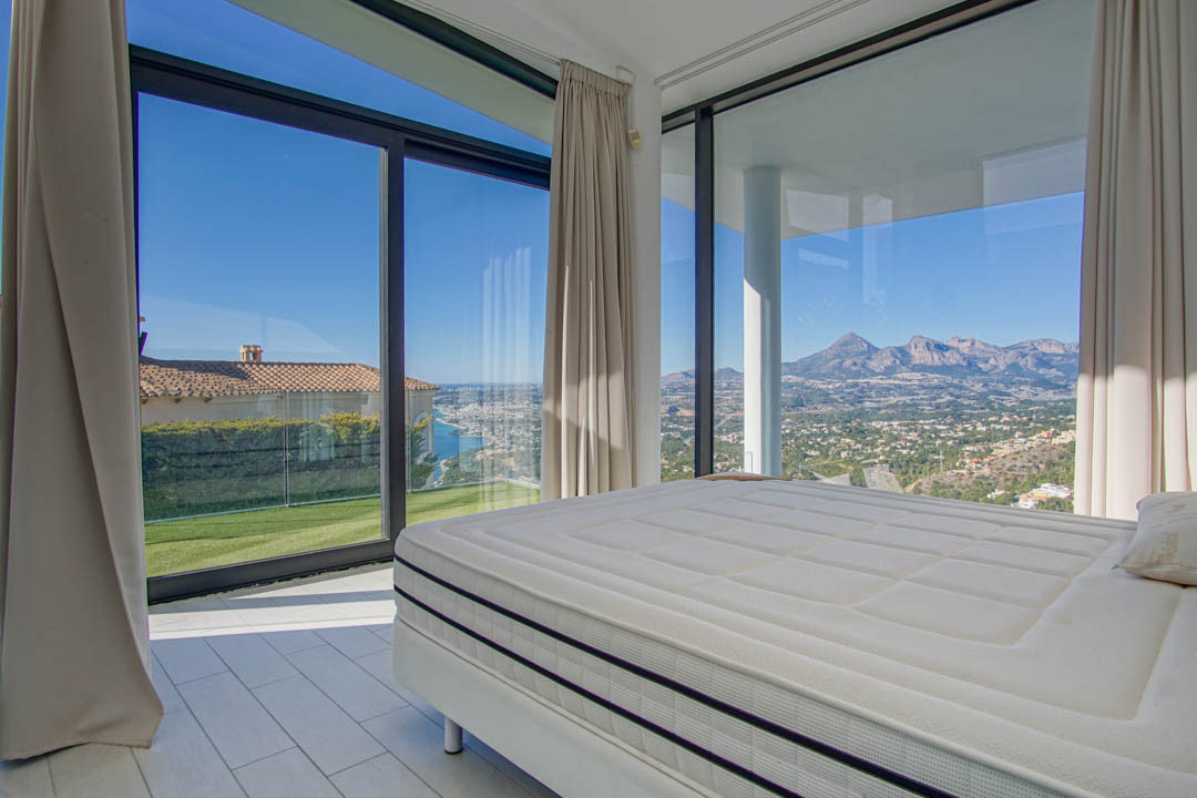 Modern bedroom with a large bed and expansive windows offering panoramic views. The vista includes a distant sea, a green lawn, and rugged mountains under a clear sky.