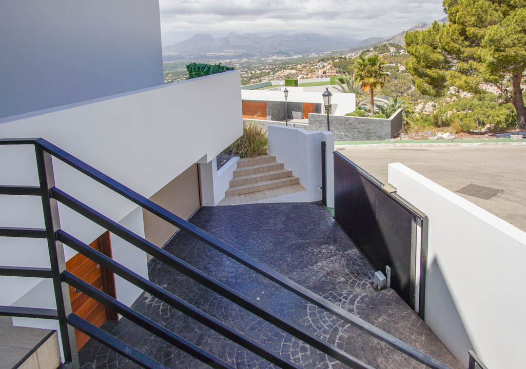 Modern exterior staircase with black railings descends to a textured dark gray driveway, featuring an an automatic gate, garage entrance, and panoramic mountain views.