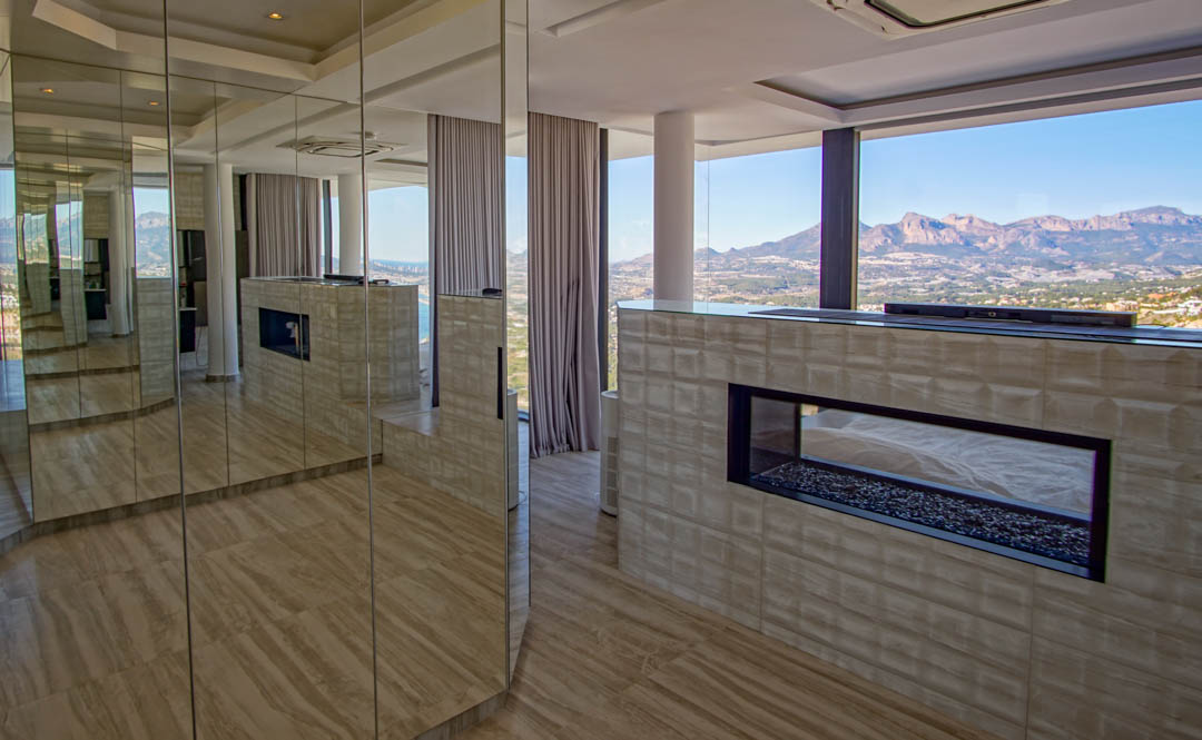 Modern interior featuring a transparent gas fireplace with pebble base, set within a textured wall. Mirrored panels reflect the room and the expansive mountain and sea views through floor-to-ceiling windows.