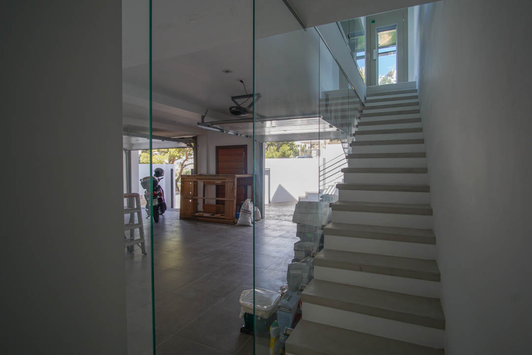 Modern interior staircase with glass balustrade and light-colored treads, adjacent to a tiled garage with an open door and natural light.