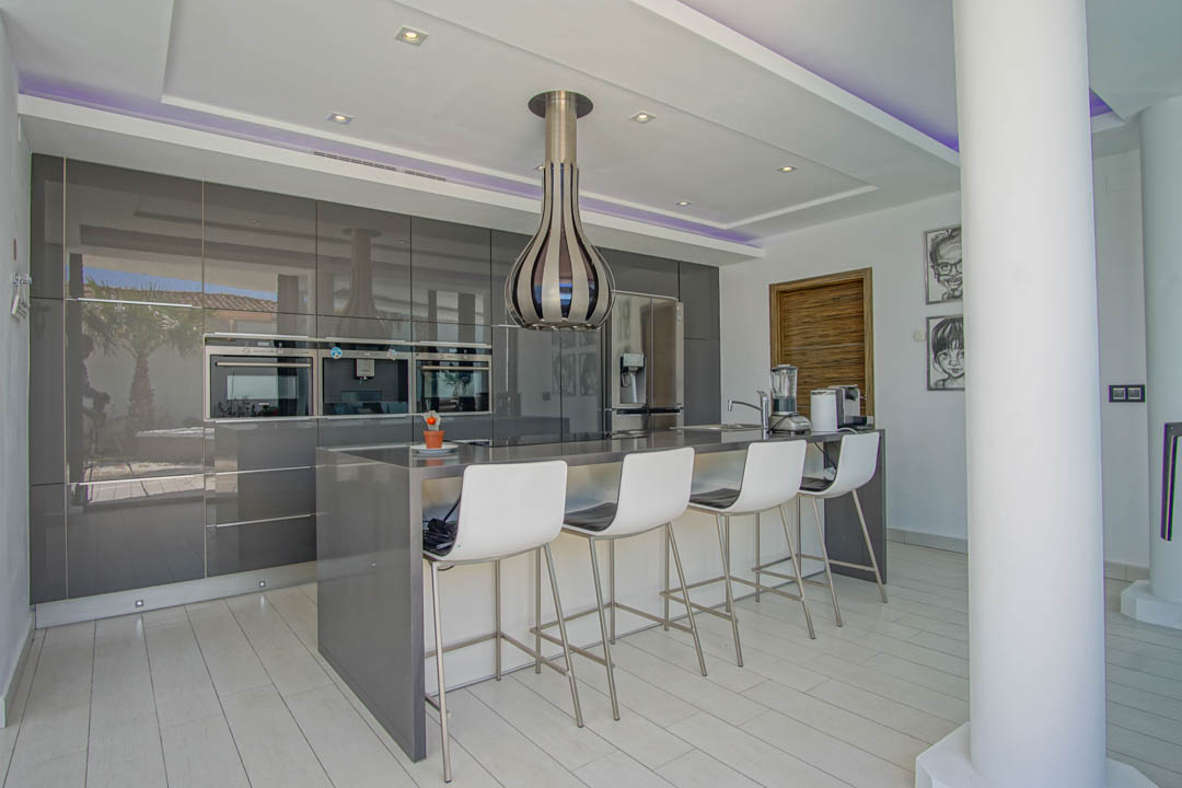 Modern kitchen featuring high-gloss grey cabinetry, integrated ovens, and a central island with seating for four. Recessed and LED strip lighting illuminate the space.