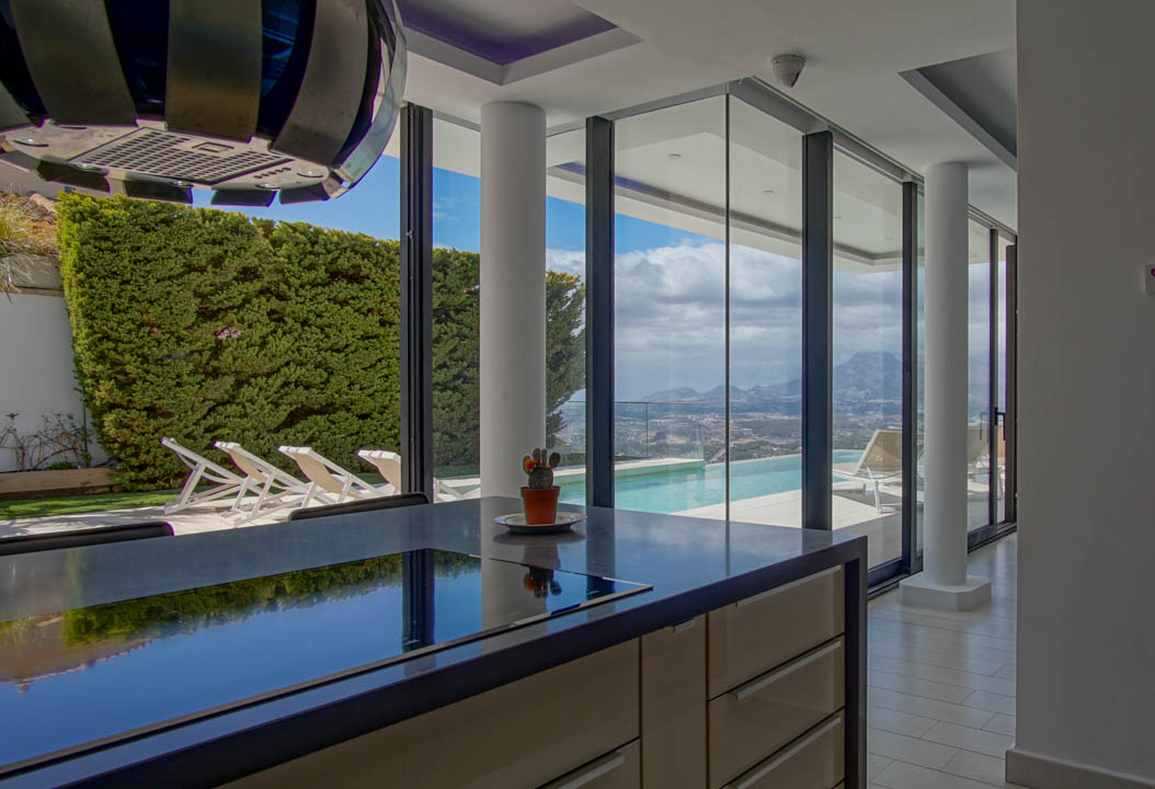 Modern kitchen island featuring a dark, reflective countertop with an integrated hob. Floor-to-ceiling glass doors open to an infinity pool and panoramic mountain views.