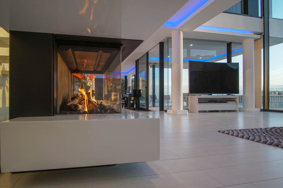 Modern living space featuring a glass-enclosed gas fireplace with a white base. Large windows and blue accent lighting complement the design.