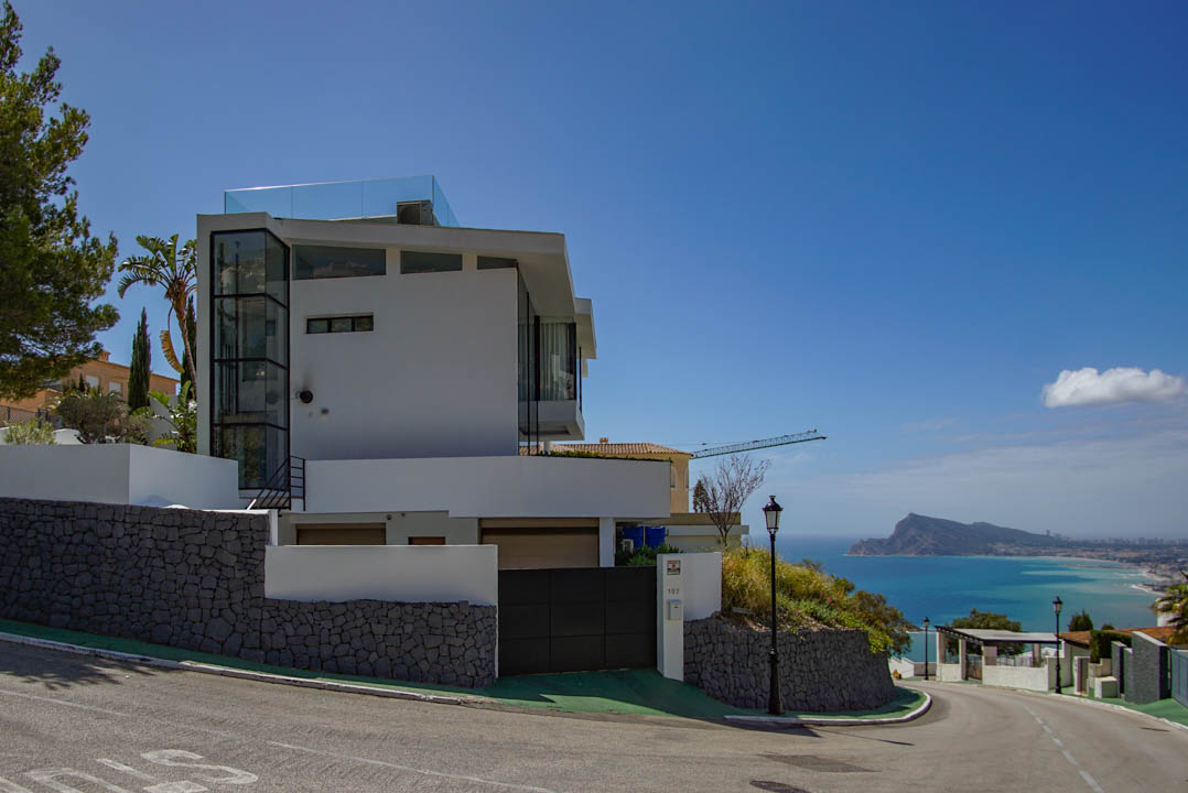 Modern white villa exterior with a prominent glass elevator shaft and rooftop terrace railing. Features stone retaining walls, an automatic gate, and panoramic sea views of the coastline.