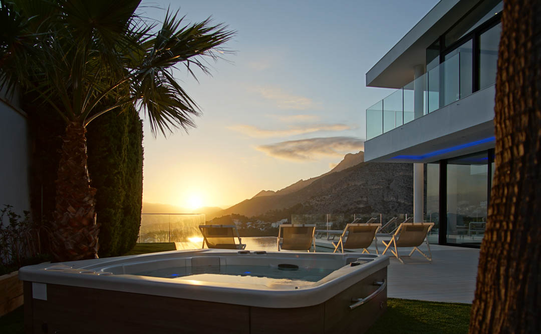Outdoor jacuzzi with wood-like finish, set on green grass, facing a modern villa with glass balconies. Features sunset mountain and sea views.