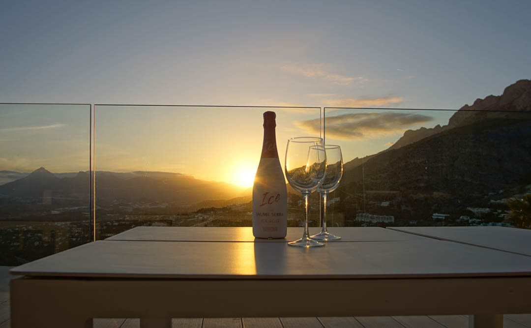 Outdoor table on a terrace with glass balustrade, featuring a rosé cava bottle and two glasses. Sunset over mountains and valley, with warm reflections.