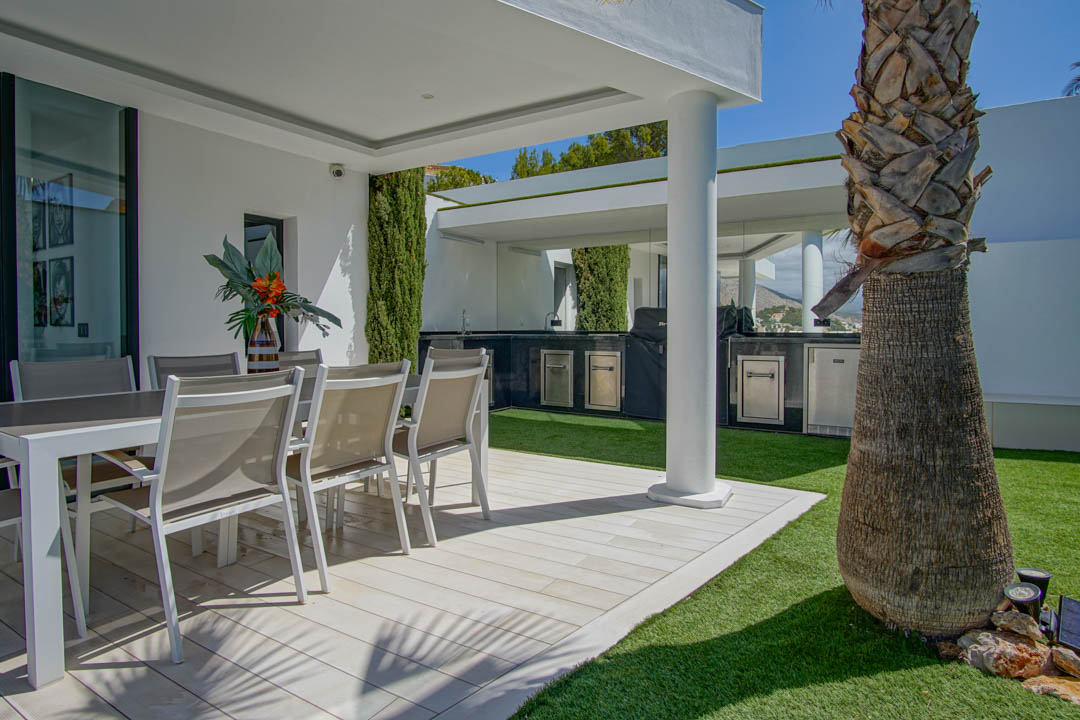 Outdoor terrace with al fresco dining on light plank-style tiling, bordered by artificial grass. Features a covered outdoor kitchen with BBQ and stainless steel appliances, beside a large palm tree.