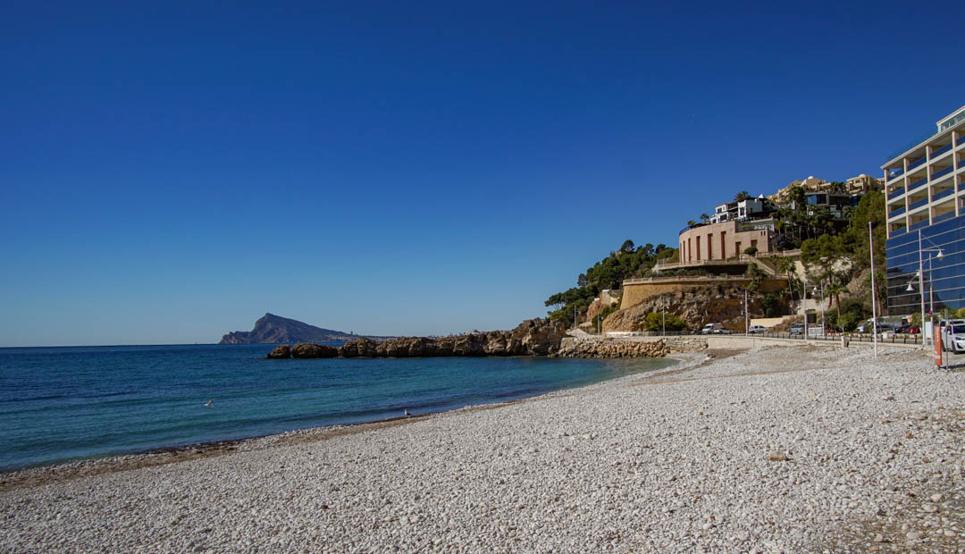 Pebble beach with clear turquoise waters and a rocky coastline. Modern buildings with reflective facades and a rounded structure line the adjacent hillside, all under a bright blue sky.