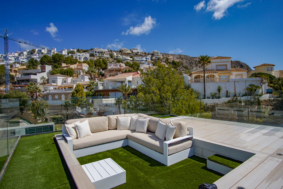 Rooftop lounge area featuring an L-shaped sofa on artificial turf, complemented by a white coffee table and glass railings. Offers clear views of the terraced hillside with residential homes.