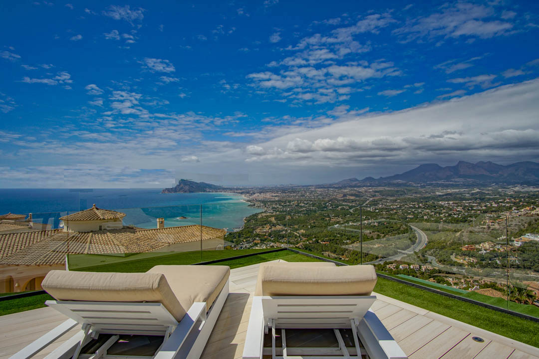 Rooftop terrace with two white sun loungers on a light wooden deck and artificial grass, offering panoramic views of the Mediterranean Sea, Altea coastline, and distant mountains.