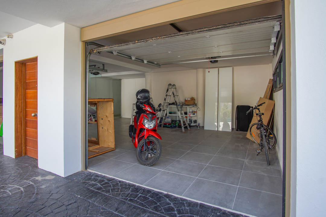 Spacious garage featuring grey tiled flooring, a partially open sectional door, and a distinct wooden entrance door on the left. Inside, a red scooter and bicycle are visible.