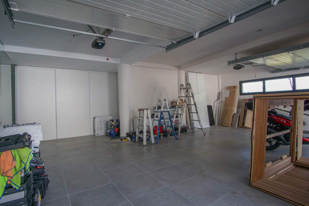 Spacious garage featuring grey tiled flooring and white walls. Equipped with two automatic door mechanisms, high horizontal windows, and white sliding storage doors.