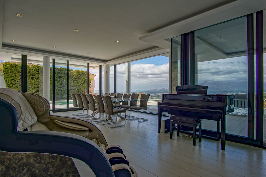 Spacious living and dining area featuring a glass table for ten, a grand piano, and a massage chair. Expansive windows provide panoramic mountain and landscape views.