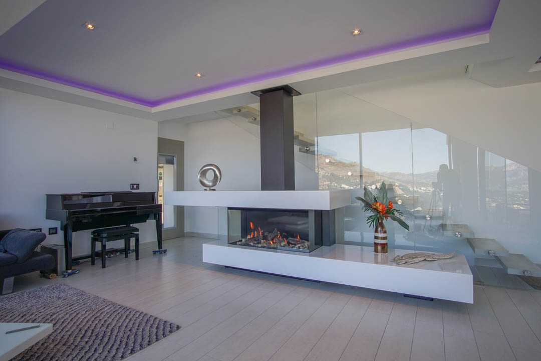 The elegant living area features a modern transparent gas fireplace, a grand piano, and a sleek glass staircase, all set against mountain views.