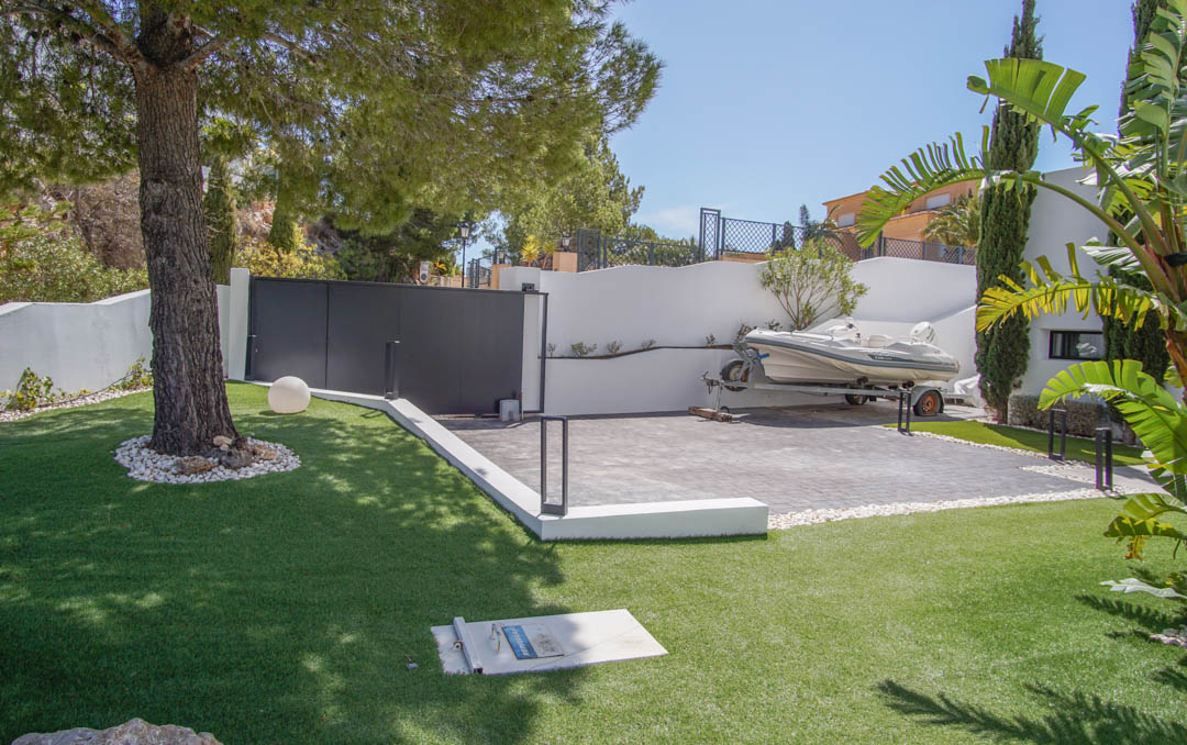 The main entrance features a dark automatic gate and a paved parking area, currently accommodating a boat on a trailer. Artificial grass and a mature pine tree define the foreground.