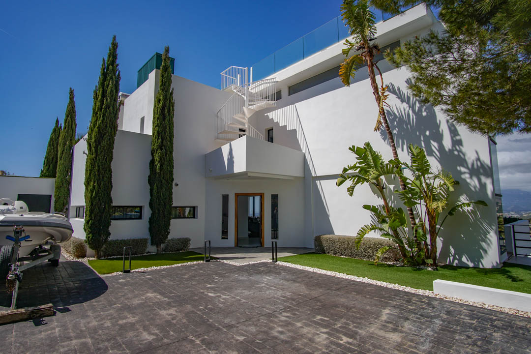 The modern white villa exterior features a paved driveway, tall cypress trees, and a distinctive spiral staircase leading to an upper terrace with glass balustrade.