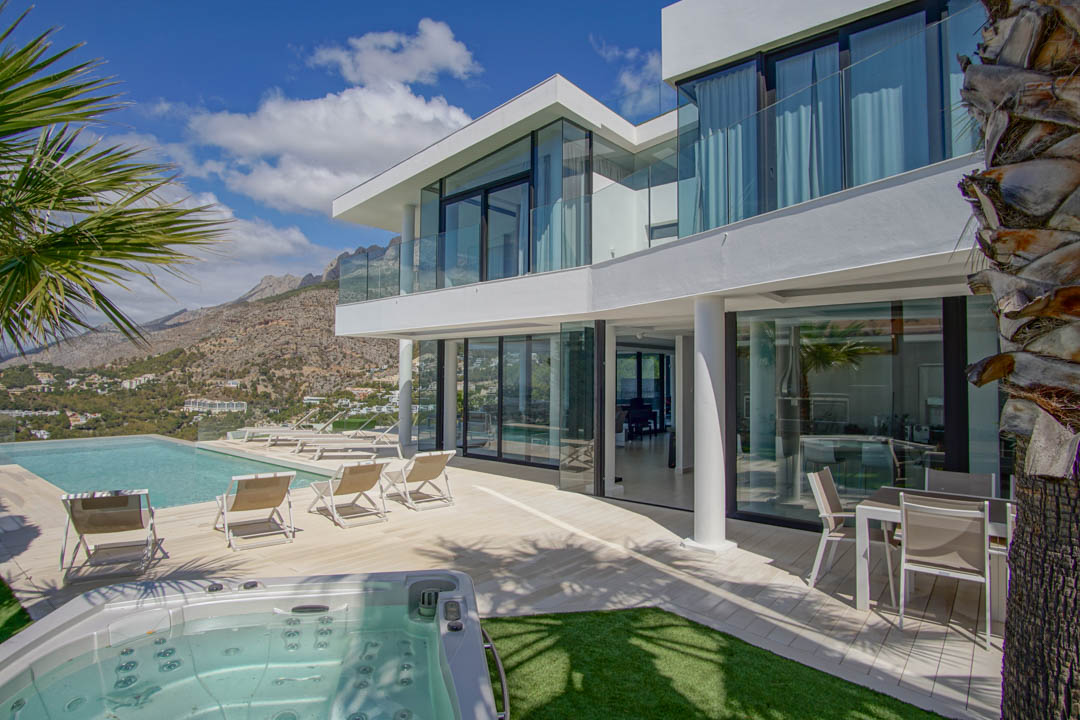 The modern white villa exterior showcases an infinity pool, separate jacuzzi, and a spacious terrace with loungers, all framed by dramatic mountain vistas under a clear sky.