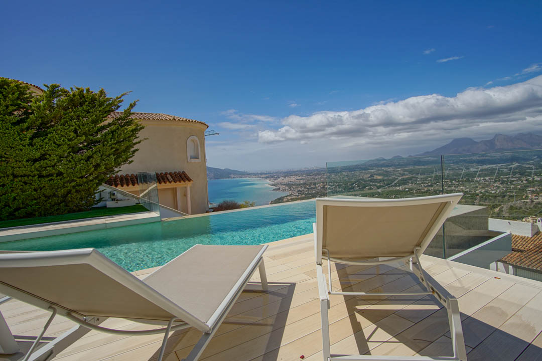 The outdoor terrace features an infinity pool overlooking the coastline and distant mountains. Two sun loungers rest on light decking, framed by a glass railing and a traditional villa facade.