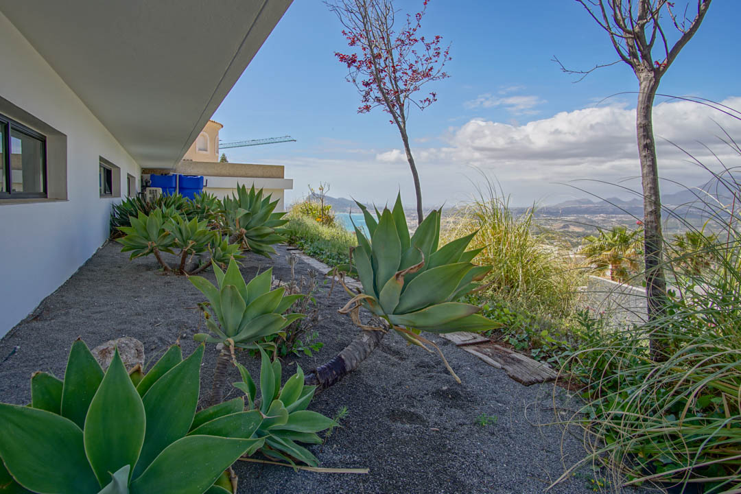 The side garden showcases large agave plants on dark gravel, bordering a white facade with dark-framed windows. It offers panoramic views of the sea and distant mountains.
