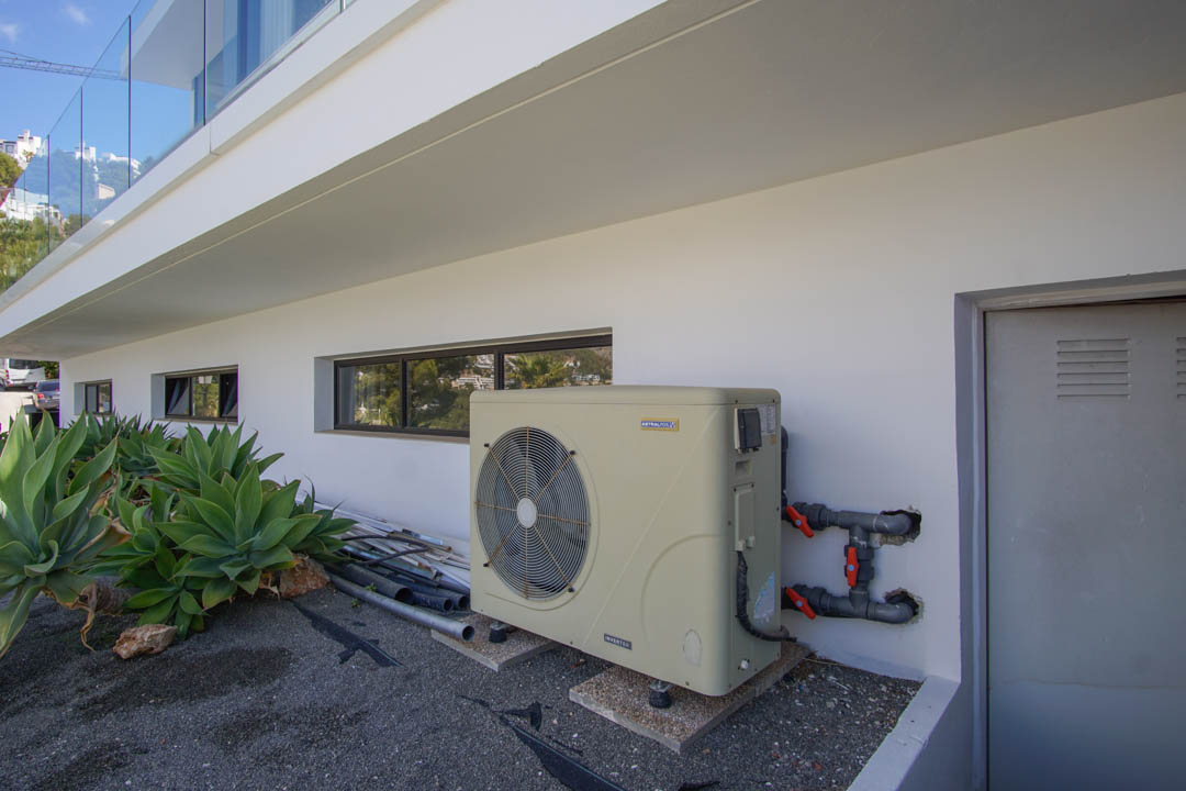 The villa's exterior technical area features an AstralPool inverter heat pump, horizontal windows, and a utility door. Agave plants complement the modern white facade.