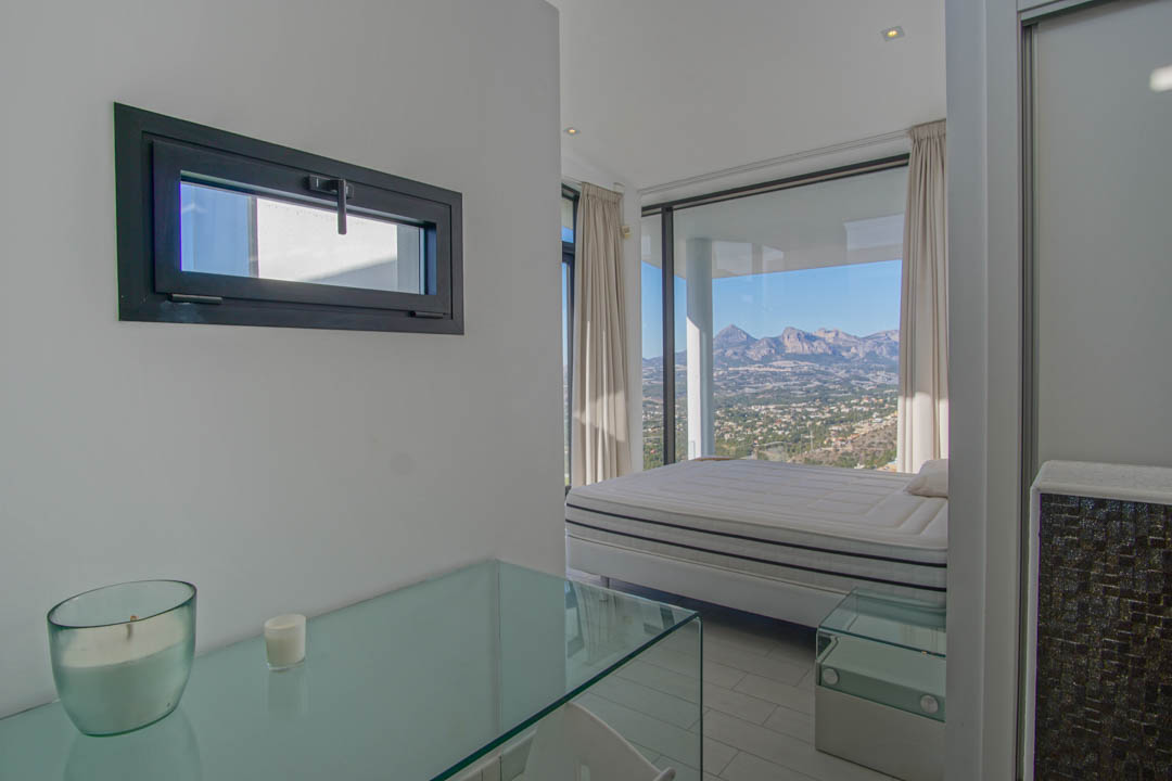 This bright bedroom features a bed positioned before expansive floor-to-ceiling windows, offering panoramic mountain and valley views. A glass console and a dark-framed horizontal window are also visible.