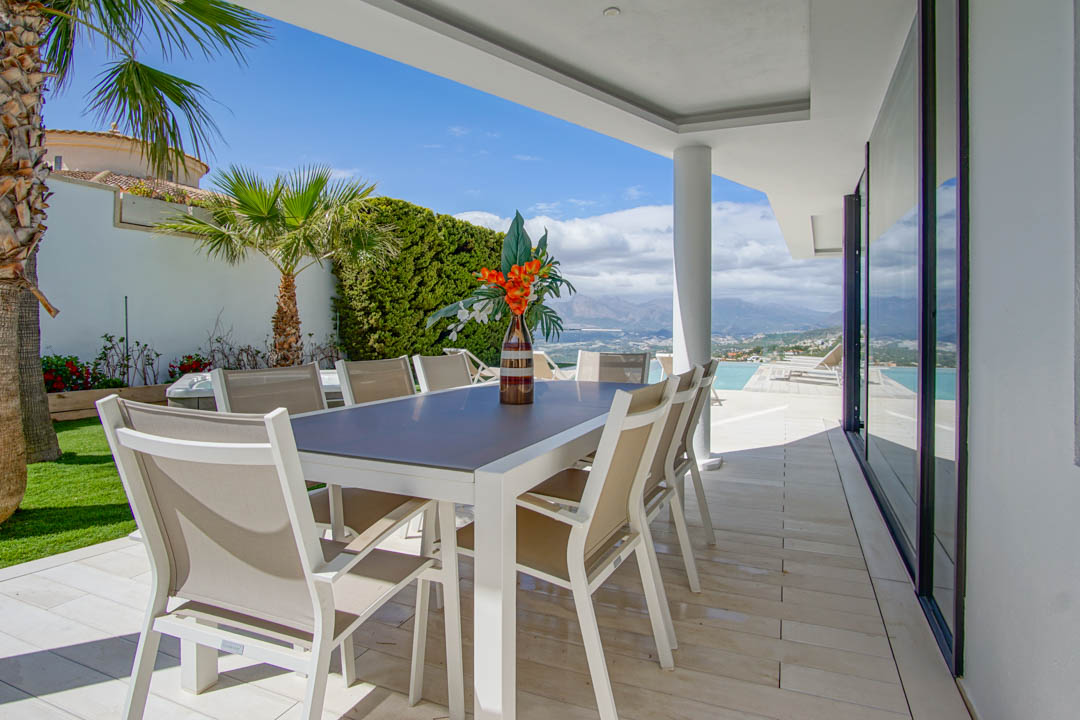 This covered outdoor dining terrace features a modern white table with eight beige chairs, set on light tiles. It offers expansive views of the distant mountains and sea, adjacent to the infinity pool.