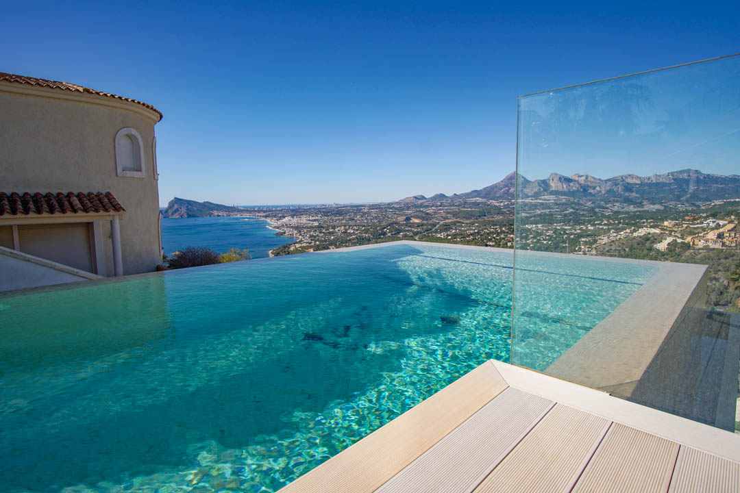 This infinity pool features clear blue water, light decking, and a glass barrier, offering panoramic views of the sea, coastline, and distant mountains under a clear sky.