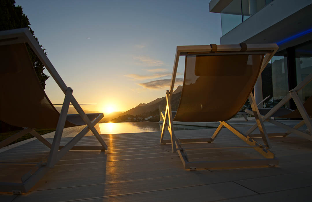 Two modern lounge chairs on light decking overlook an infinity pool, reflecting a golden sunset over distant mountains.