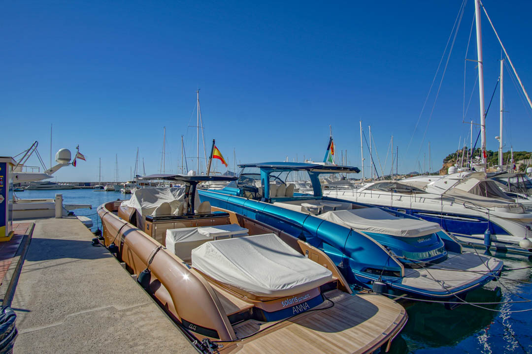 Two modern powerboats, one bronze with a teak deck and another metallic blue, docked in a marina under a clear sky. Spanish flags are visible among the background yachts.