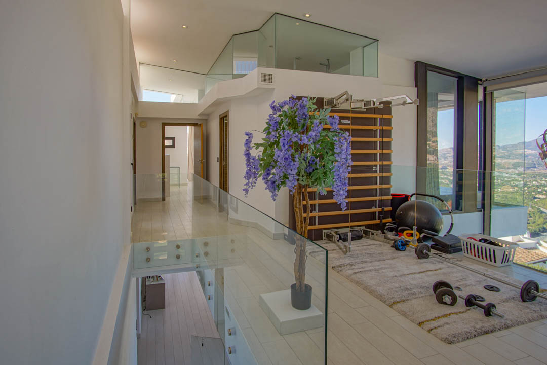 Upper floor gym area featuring modern glass railings, light wood-effect flooring, and a wall-mounted exercise ladder. Panoramic views of the surrounding landscape are visible through expansive floor-to-ceiling windows.