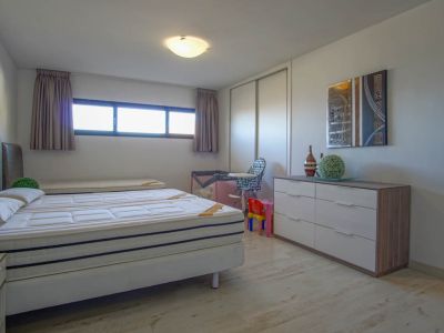 A bright bedroom featuring a bed with two mattresses, light wood-look flooring, and a long horizontal window. The room includes a white sliding wardrobe and a modern dresser with an abstract artwork above.