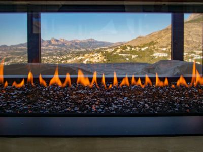 A modern gas fireplace with active flames over dark glass media, set against a panoramic window revealing distant mountains and a valley landscape.