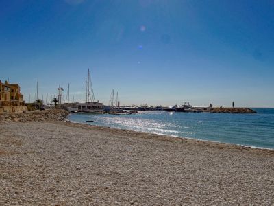 A wide pebble beach stretches towards a marina filled with yachts, flanked by traditional coastal buildings under a bright, clear blue sky.