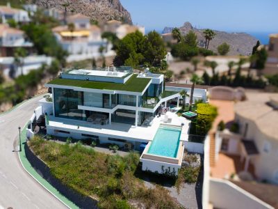 Aerial view of a contemporary white villa with an infinity pool, extensive glass facades, a green roof, and a separate jacuzzi, offering mountain and sea views.