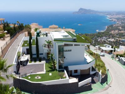 Aerial view of a modern white villa featuring multi-level architecture, green flat roofs, and an external glass elevator. The property includes a private driveway and offers panoramic sea and coastal views.