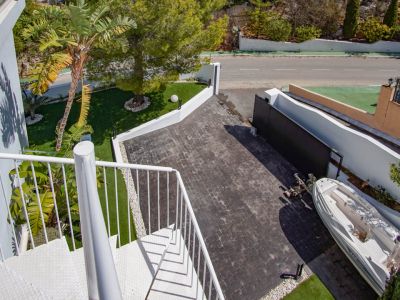 Aerial view of the main entrance featuring a white spiral staircase, a dark paved driveway, and artificial grass areas with a palm tree. An automatic gate and parking space are visible.