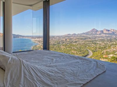 Bedroom featuring floor-to-ceiling glass, framing panoramic views of the Mediterranean Sea, a coastal town, and distant mountains. A low-profile bed with white linens faces the expansive vista.