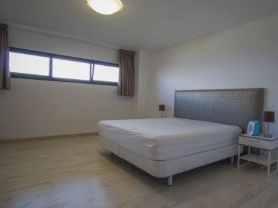 Bedroom featuring light wood-look flooring, white walls, and a bed with a textured grey headboard. A horizontal window with dark frames and curtains provides natural light.