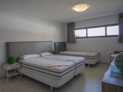 Bedroom featuring two single beds with grey patterned headboards, a third single bed in the background, light wood flooring, and a series of horizontal windows.