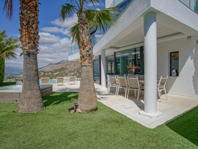 Covered outdoor dining terrace with modern furniture, adjacent to a lush artificial lawn and prominent palm trees. A jacuzzi and pool area offer mountain and distant sea views.