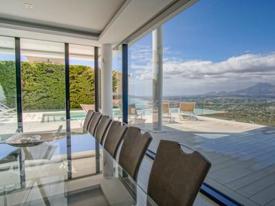 Elegant indoor dining area featuring a glass table and chairs, seamlessly connecting to an expansive terrace with an infinity pool and panoramic sea and mountain vistas.